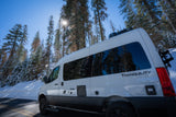 This white RV rental is parked by snow-covered trees on a sunny winter day, ready for adventure on the open road.