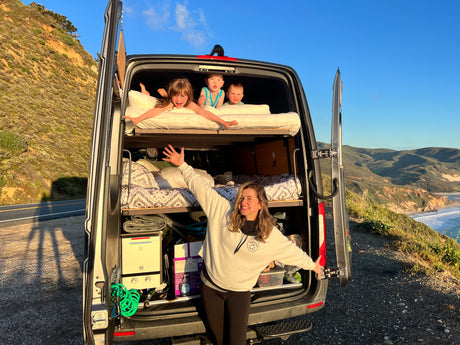 Joyful family on an RV rental adventure! Kids lounge in bunk beds in the van, mom smiles at scenic coastal views.