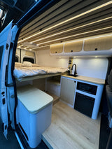 Modern RV rental interior with a raised bed, compact kitchen (microwave, fridge), overhead cabinets, and warm ceiling lights.