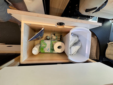 RV rental: Organized camper cabinet with essential supplies like toilet paper, wipes, and detergent, next to a trash bin.