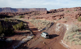 An RV rental navigates a remote dirt road through arid red rock canyons. Ideal for adventurous desert exploration.