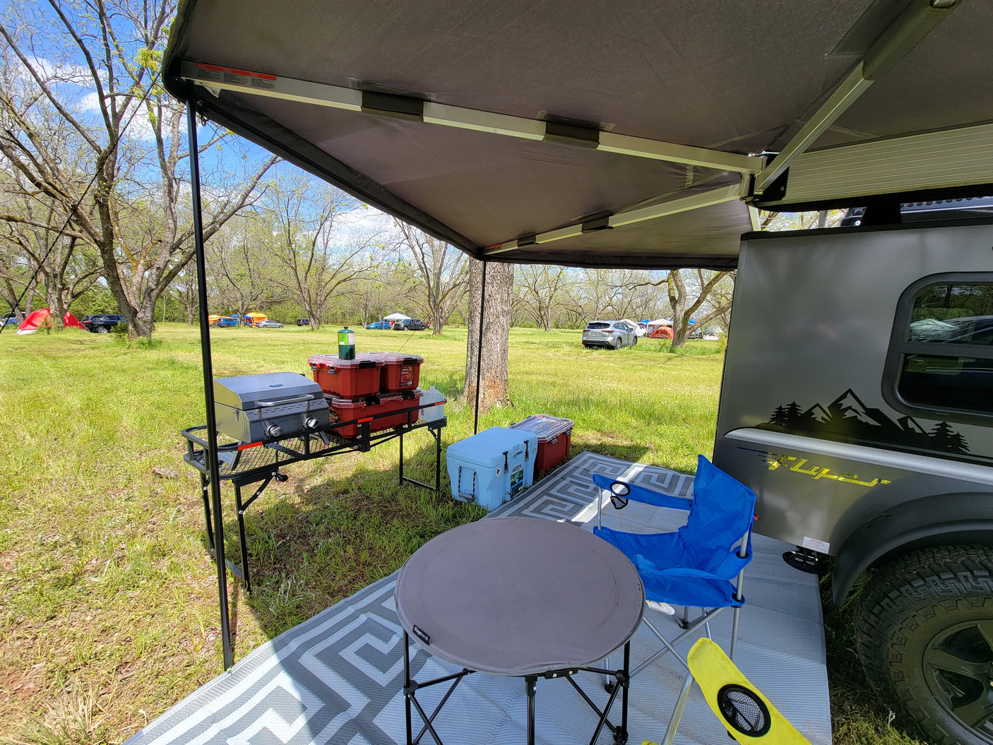 Scenic RV rental campsite features a small camper with an extended awning, portable grill, coolers, and outdoor seating.