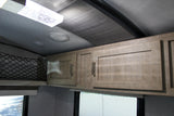 RV rental interior featuring light wood overhead cabinets, net storage, ceiling lights, speakers, and window blinds.