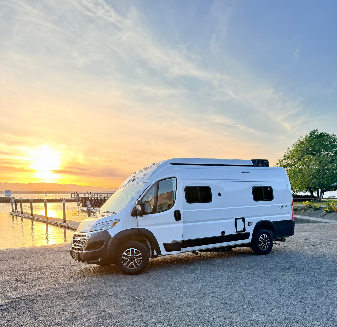 Experience freedom! A white Winnebago Solis rv rental parked by a peaceful waterfront, bathed in sunset's golden light.