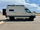 This light-colored camper van, equipped with a roof rack & side steps, ideal for RV rental, is parked on asphalt under a cloudy sky.