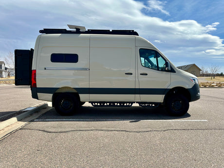 This light-colored camper van, equipped with a roof rack & side steps, ideal for RV rental, is parked on asphalt under a cloudy sky.