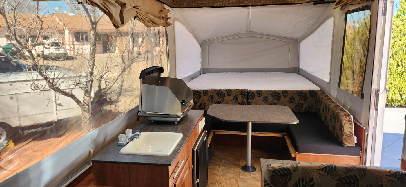 Pop-up camper RV rental interior featuring a large bed, dinette with table, and compact kitchen area with sink and stove. — photo 2