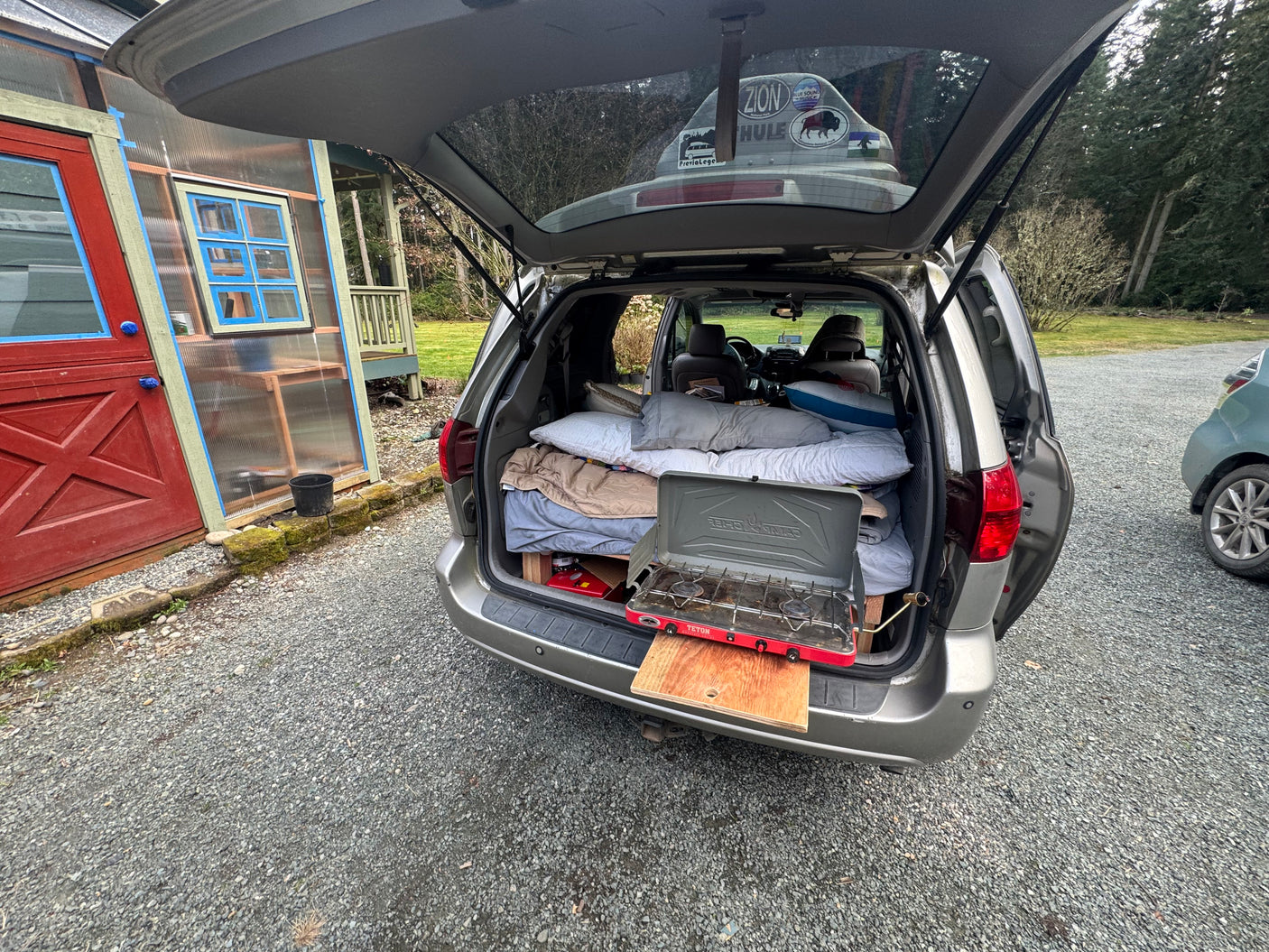 A cozy minivan RV rental setup: a bed with pillows, and a Teton camping stove on a pull-out. Ready for your next road trip!