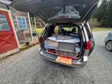 A cozy minivan RV rental setup: a bed with pillows, and a Teton camping stove on a pull-out. Ready for your next road trip!