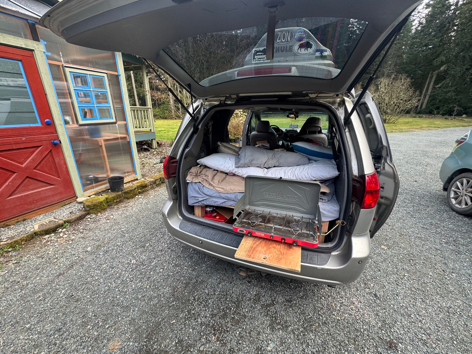 A cozy minivan RV rental setup: a bed with pillows, and a Teton camping stove on a pull-out. Ready for your next road trip!