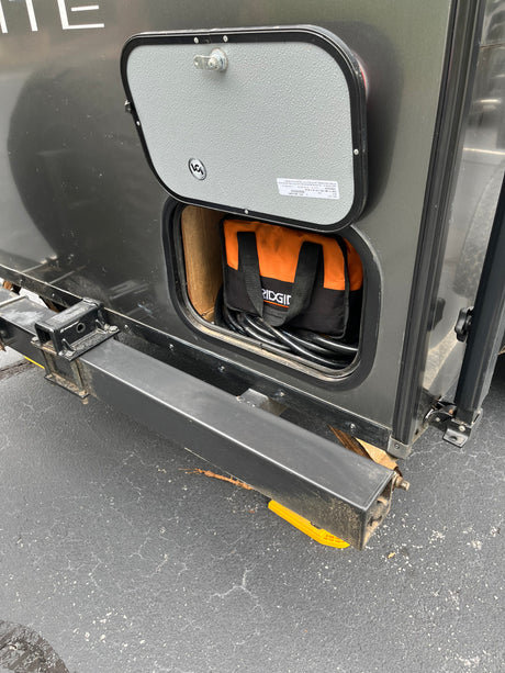 RV rental utility: Open storage hatch on a dark gray RV. Inside are coiled cables and a RIDGID bag. Rear bumper, yellow leveling block.