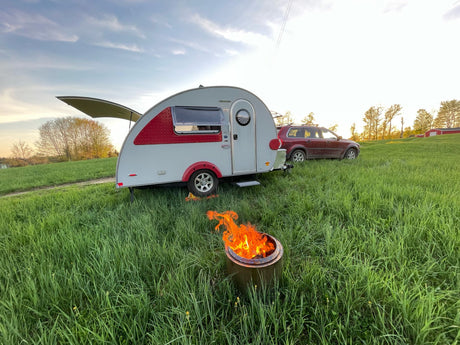 Teardrop RV rental & SUV camping in a grassy field with a campfire at sunset.