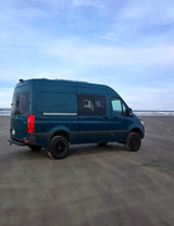 This teal camper van RV rental sits on a sandy beach next to the ocean waves under a blue sky, ready for adventure.