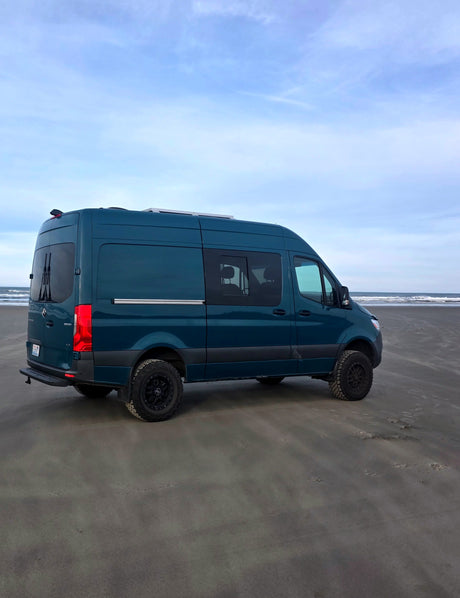 This teal camper van RV rental sits on a sandy beach next to the ocean waves under a blue sky, ready for adventure.