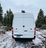 Rear view of a white Mercedes Sprinter rv rental van in a snowy forest with pine trees under an overcast sky.