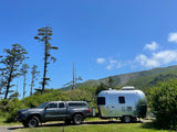 This RV rental combo features a grey truck towing a shiny Airstream travel trailer, parked in a sunny, tree-lined campsite.