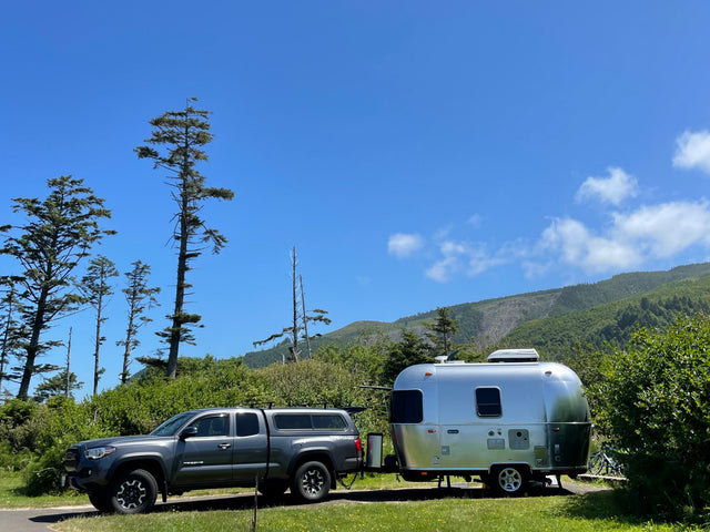 This RV rental combo features a grey truck towing a shiny Airstream travel trailer, parked in a sunny, tree-lined campsite.