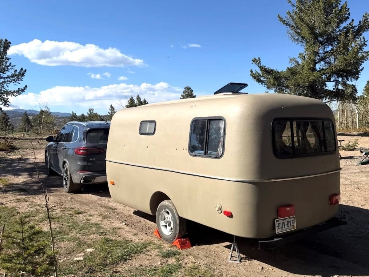 Tan retro rv rental and SUV parked on a rustic campsite among pine trees with mountains in the distance under a blue sky.|This olive-colored RV rental camper is ready for adventure — photo 1