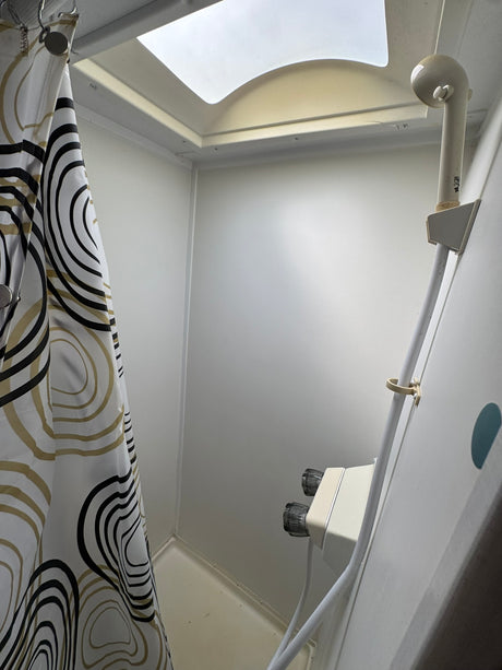 Inside an RV rental shower: white walls, a bold patterned curtain on the left, shower head on the right, and a ceiling skylight.