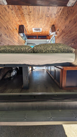 RV rental camper van interior: features wood paneling, an elevated bed with patterned pillows, and under-bed storage.