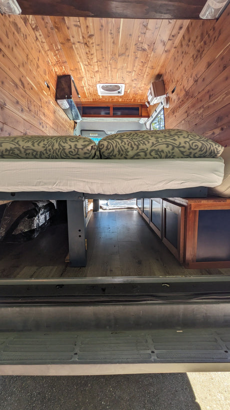 RV rental camper van interior: features wood paneling, an elevated bed with patterned pillows, and under-bed storage.