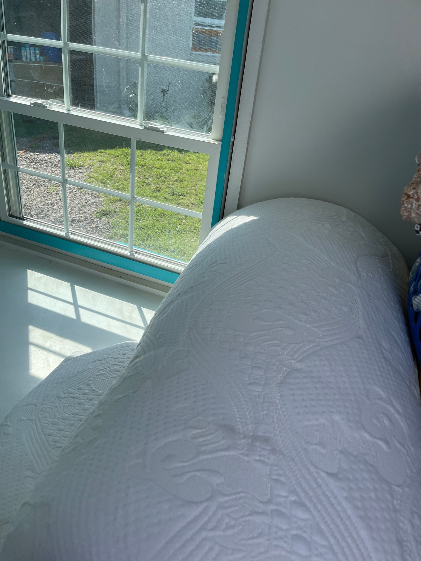 RV rental interior featuring a window view of grass and gravel, with a textured white bed. Sunlight casts shadows.