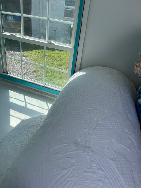 RV rental interior featuring a window view of grass and gravel, with a textured white bed. Sunlight casts shadows.