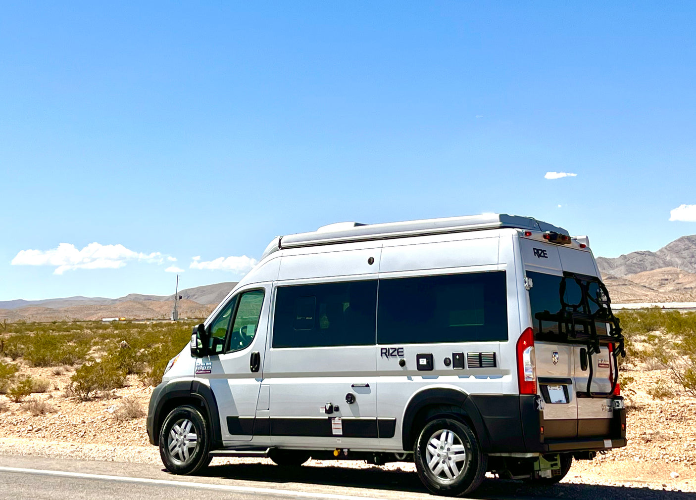 A white RIZE camper van, perfect for an RV rental, is parked on a desert roadside with distant mountains under a blue sky.