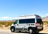 A white RIZE camper van, perfect for an RV rental, is parked on a desert roadside with distant mountains under a blue sky.