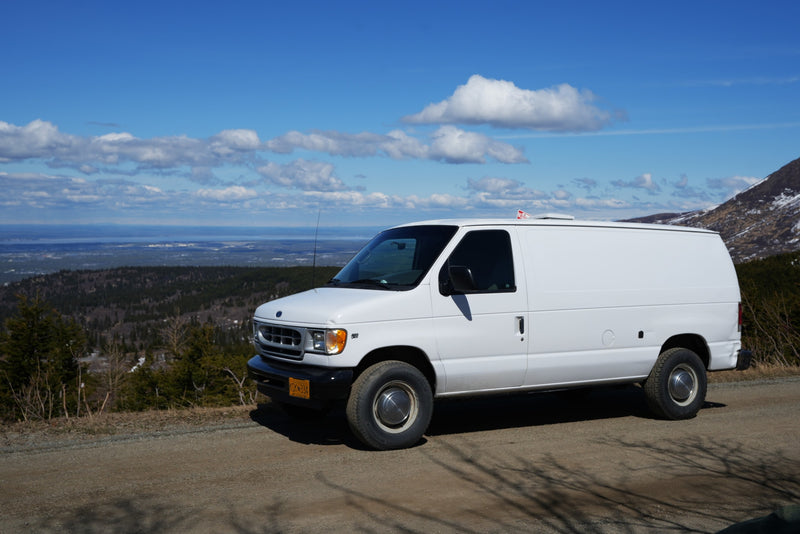 A white RV rental van is parked on an Alaskan mountain road, offering stunning views of forests and an inlet under a blue sky. — photo 11