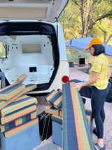 Preparing an RV rental! Woman loads striped cushions into the small travel trailer's open hatch for a comfy trip.