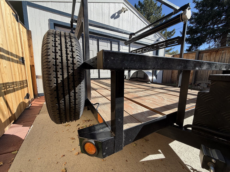 Passenger-side view along the trailer frame rail showing the spare tire mounted at the front, the amber side marker light, the wood-plank deck and the overhead rack uprights — photo 6