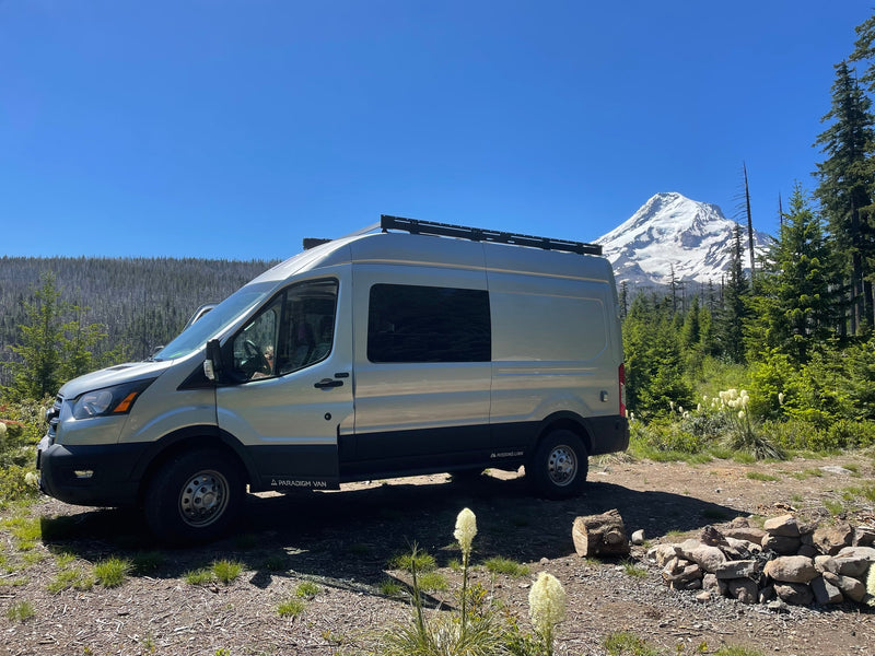 RV rental: A grey Ford Transit camper van at a sunny forest campsite, snowy mountain & fire pit. — photo 2