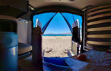 RV rental camper van interior. View out open rear doors to a sunny beach, blue lake, and distant mountains. Bed and fruit visible.
