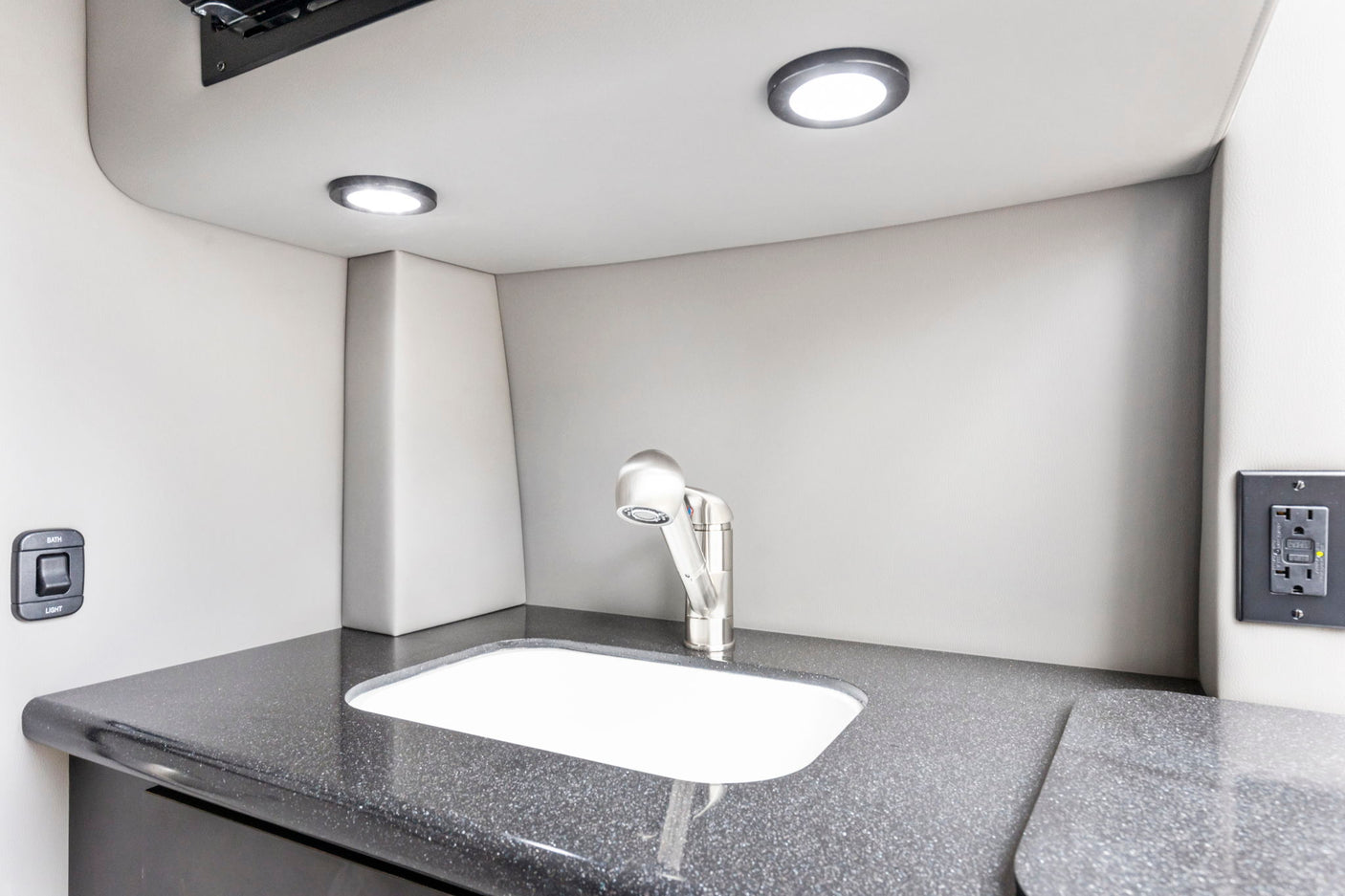 RV rental bathroom/kitchenette: compact sink, modern faucet, dark counter, and bright LED lights. Features switches & outlet.