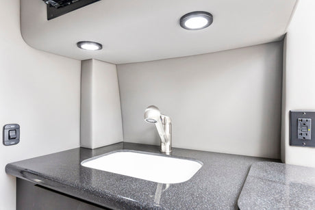 RV rental bathroom/kitchenette: compact sink, modern faucet, dark counter, and bright LED lights. Features switches & outlet.