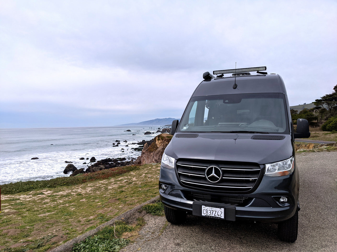 Dark gray Mercedes-Benz camper van (REC VAN) on a cloudy day overlooking a rocky California coast, ideal for an RV rental.