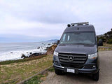 Dark gray Mercedes-Benz camper van (REC VAN) on a cloudy day overlooking a rocky California coast, ideal for an RV rental.