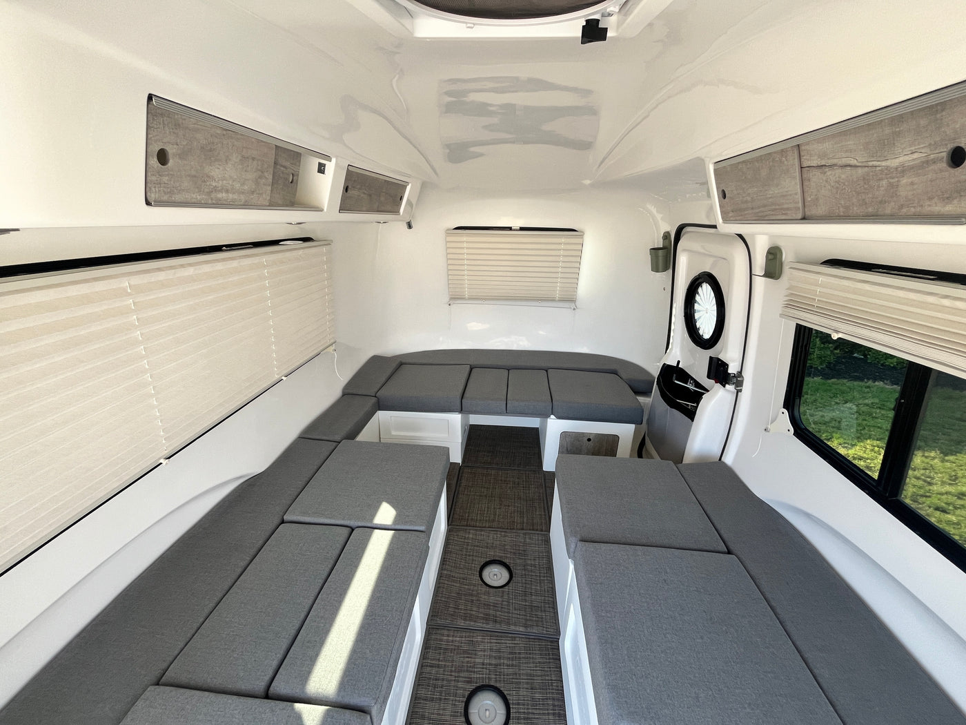 Clean RV rental interior: U-shaped grey seating converts to beds, with overhead storage, blinds, and dark floor for travel. — photo 14