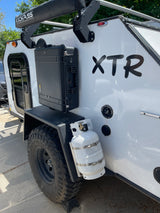 White off-road RV rental camper with "XTR" decal, a robust storage case, propane tank, and a "KICKASS" awning.