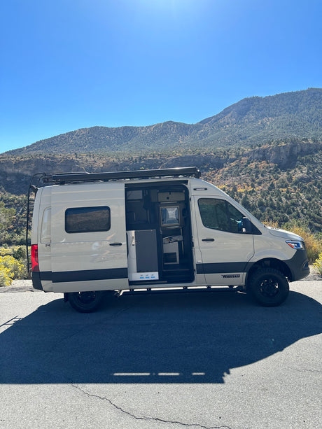 RV rental camper van parked on a mountain road with its side door open, revealing the interior under a clear blue sky.