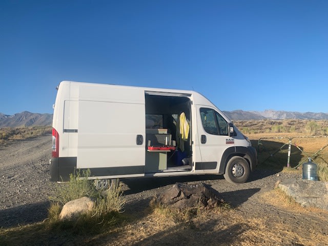 An rv rental camper van parked on a dirt road, side door open to reveal a kitchen setup, with mountains in the distance under a blue sky. — photo 7