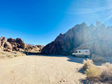 White RV rental parked among towering desert rock formations on a sunny day in a vast, sandy desert landscape.