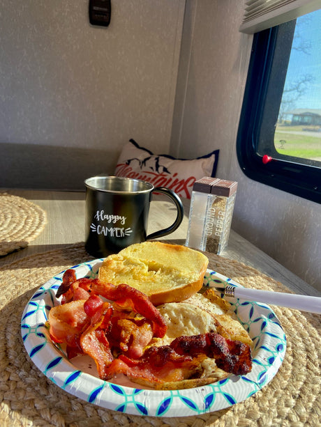 A cozy RV rental interior with a sunny breakfast: bacon, eggs, and toast. "Happy Camper" mug & pepper shaker on the table.