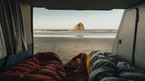 Inside an RV rental, two sleeping bags face a serene beach with a large sea stack in the ocean.