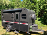 A sleek black teardrop camping trailer, ideal for an RV rental adventure, features off-road tires, roof rack, and side awning.