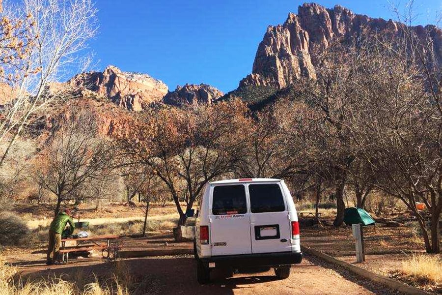  surrounded by autumn trees and red rock mountains. Ideal for an RV rental adventure.|White campervan (RV rental) at a stunning desert campsite with majestic red rock mountains and bare trees. Perfect for adventure!|White RV rental van at a scenic park campsite — photo 2