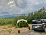 A green tent and grey car parked at a beautiful mountain campsite. Enjoy the outdoors—whether with an RV rental or a tent.