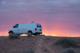 A white RV rental van sits atop a dirt hill against a vibrant purple and orange sunset sky, showcasing remote travel.