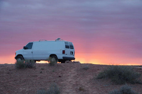 A white RV rental van sits atop a dirt hill against a vibrant purple and orange sunset sky, showcasing remote travel.